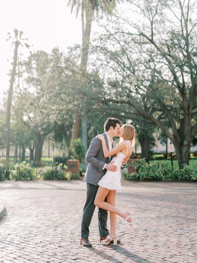 Candid engagement photo of couple kissing on brick-paved road with the sun lighting them up